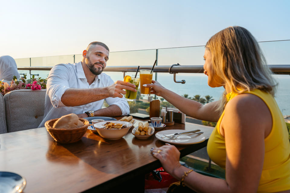 A man and woman share a toast with drinks at an outdoor restaurant by the sea. The table has various dishes and condiments