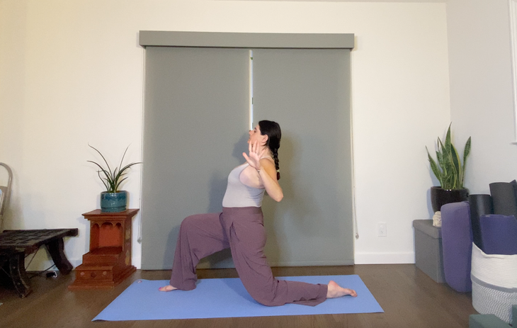 A woman wearing loose purple pants and a gray tank top practices a Low Lunge with her arms in cactus position.