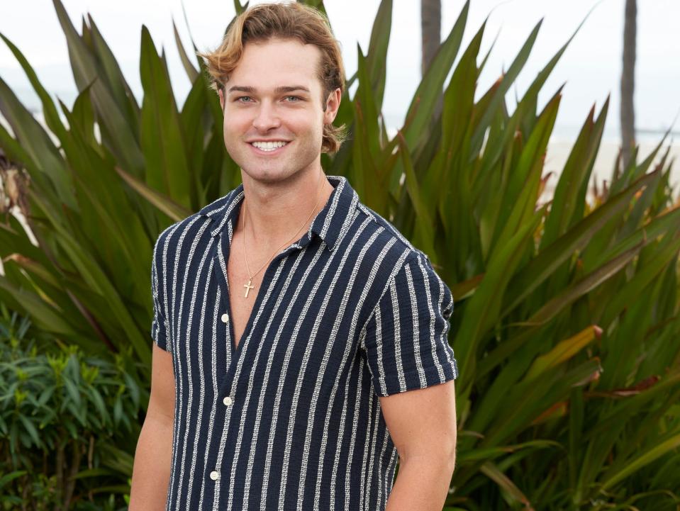 Sean McLaughlin poses in a striped shirt and white shorts in front of a tropical plant.