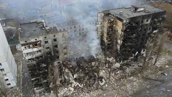 Smoke rises from a large, nine-story residential building destroyed by apparent shelling, which is completed collapsed in the middle.