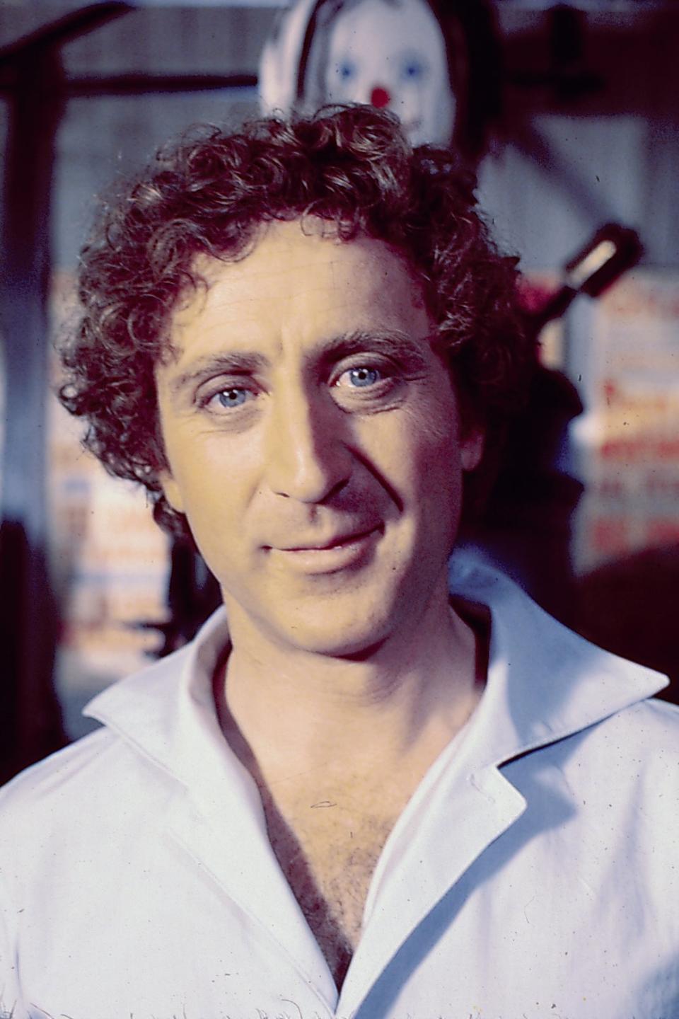 Gene Wilder with curly hair and a light shirt is smiling gently, standing indoors