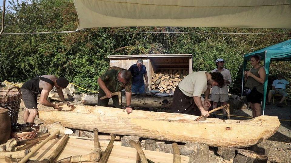 Three men using hand tools while carving a log into a boat