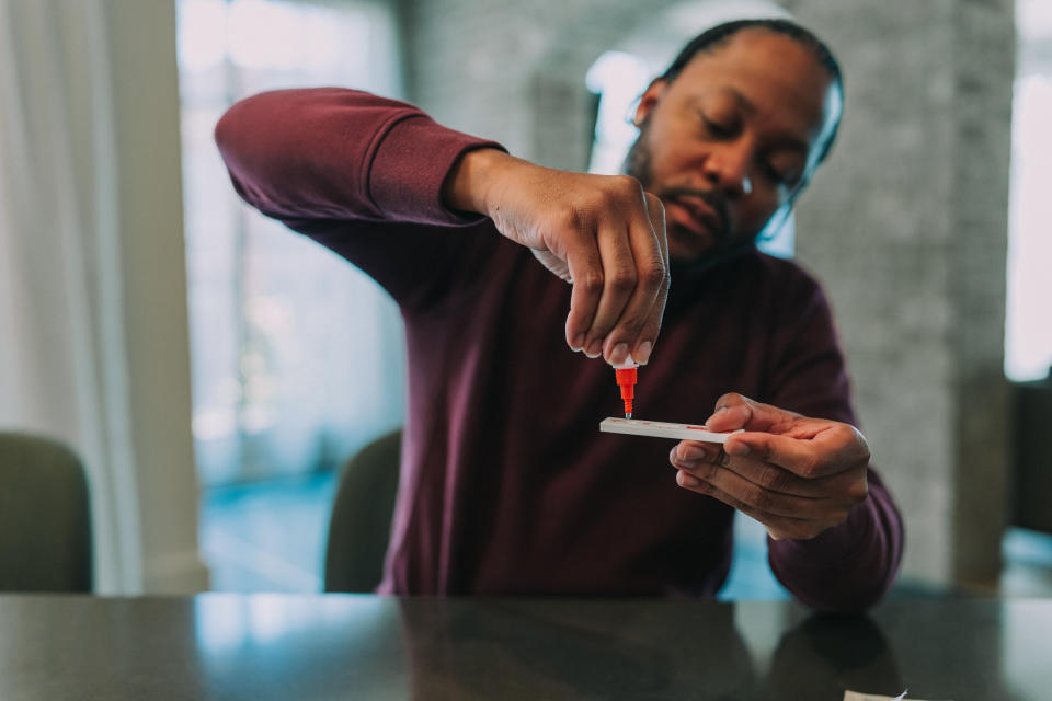 A black man sits in his kitchen and uses a dropper to place testing solution on the testing strip of a COVID-19 rapid self testing kit