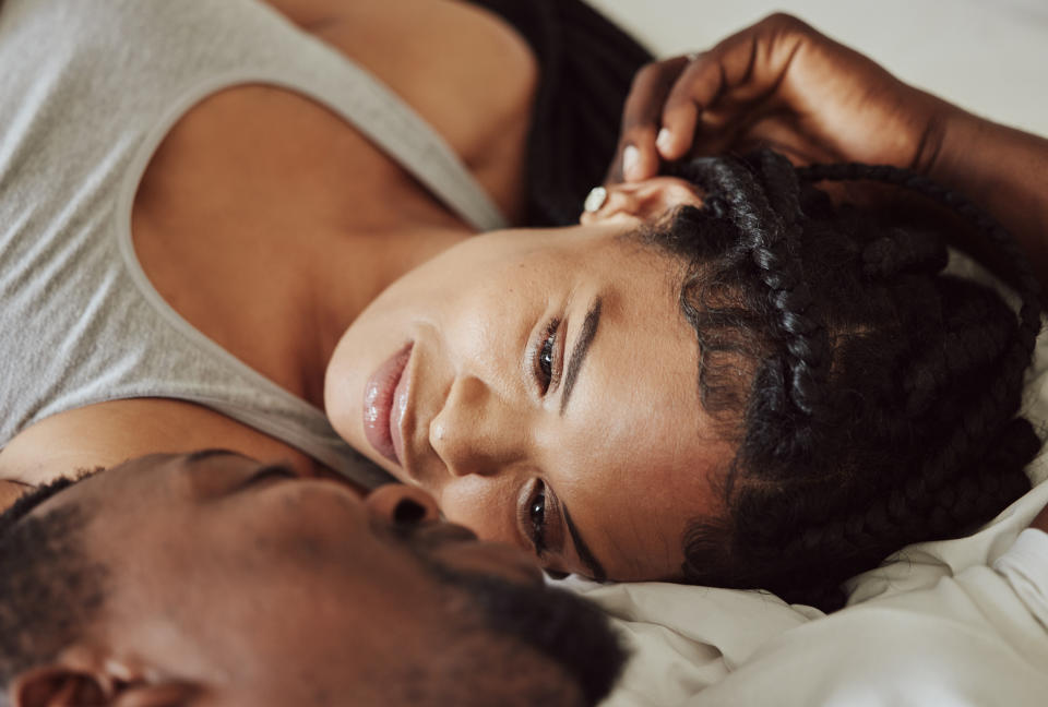 A woman with braided hair and a man lie in bed, intimately gazing into each other's eyes