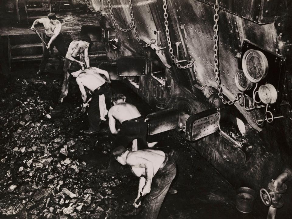 Several shirtless men shovel coal from a large pile on a ship