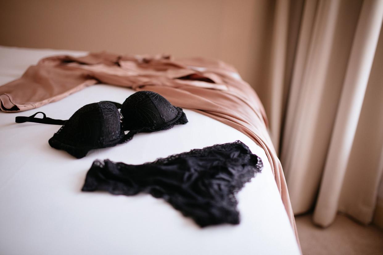 Black lace lingerie bra and panty and a beige silk robe laid on a white sheeted bed with a blurred background of a beige carpeted floor and beige window curtains