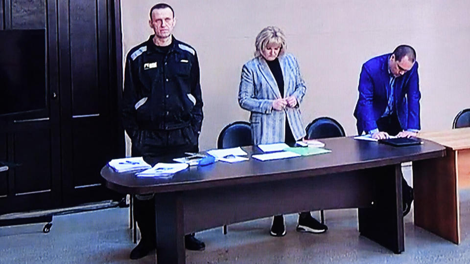 Alexei Navalny stands behind a desk with his hands in his pockets, with a woman and a man to his left, also standing, who are reviewing documents, 