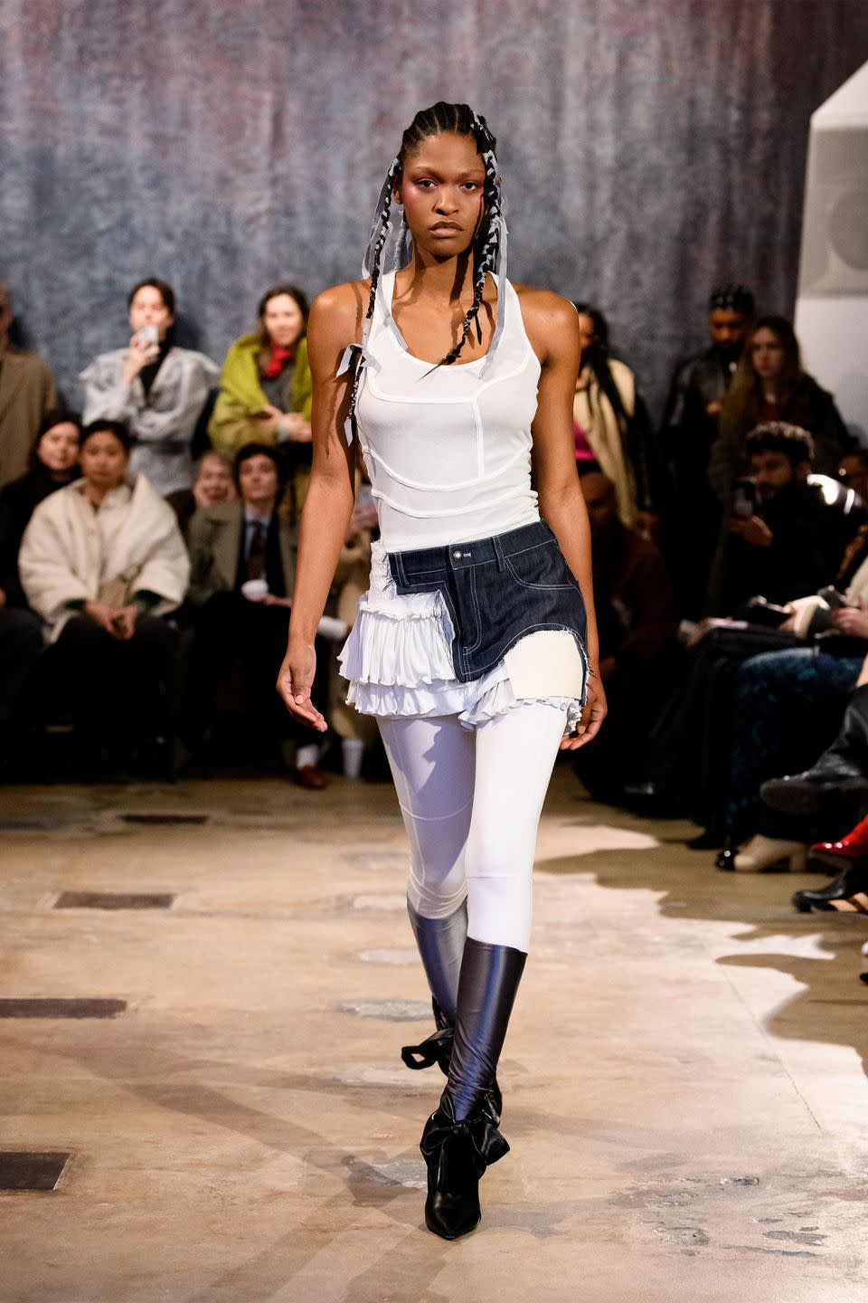 a model walks the runway in a denim miniskirt split in half with white ruffles down one side