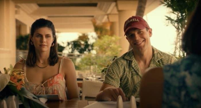 Alexandra Daddario and Jake Lacy are sitting at a dining table outdoors, talking to another person who is partially visible in the foreground