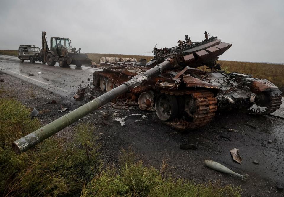 A destroyed Russian tan on a road in the foreground with a Ukrainian serviceman rides a tractor pulling a military vehicle behind it, all beneath a grey sky