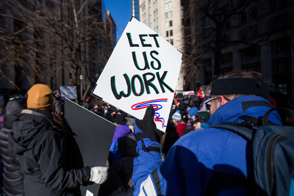 A demonstrator holds a sign that reads ‘Let Us Work’ during a protest against the government shutdown in Boston, Massachusetts, U.S., on Friday, Jan. 11, 2019. Government shutdown lawsuits over lost pay are piling up. (Photo credit: Scott Eisen/Bloomberg via Getty Images)