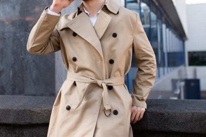 A man in a classic, tan trench coat stands outside of an office building.