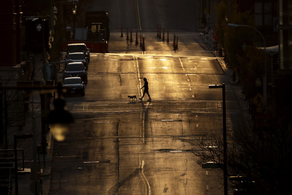 A man walks a dog on an empty downtown street as the sun sets Wednesday, April 1, 2020, in Kansas City, Mo. (AP Photo/Charlie Riedel)