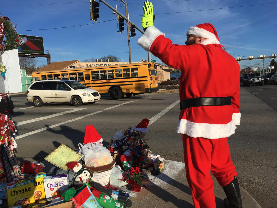 In this Dec. 18, 2018 photo, Jimmy Izbinski, wearing a Santa suit, waves to motorists passing the Christmas weed in Toledo, Ohio. The street corner weed decked out with lights and ornaments is spreading holiday goodwill. (AP Photo/John Seewer)