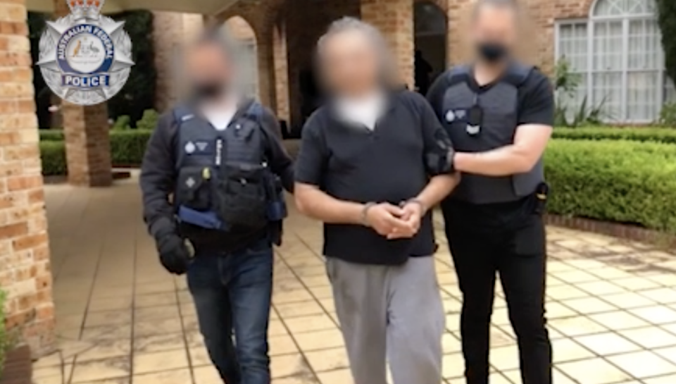 A man wearing a dark coloured shirt and grey pants being escorted out of a brick home with two police officers. Source: AFP