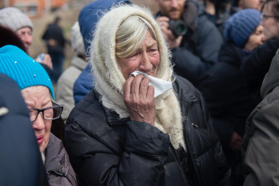 A woman, standing in a crowd. with a white scarf over her head holds a tissue to her face.