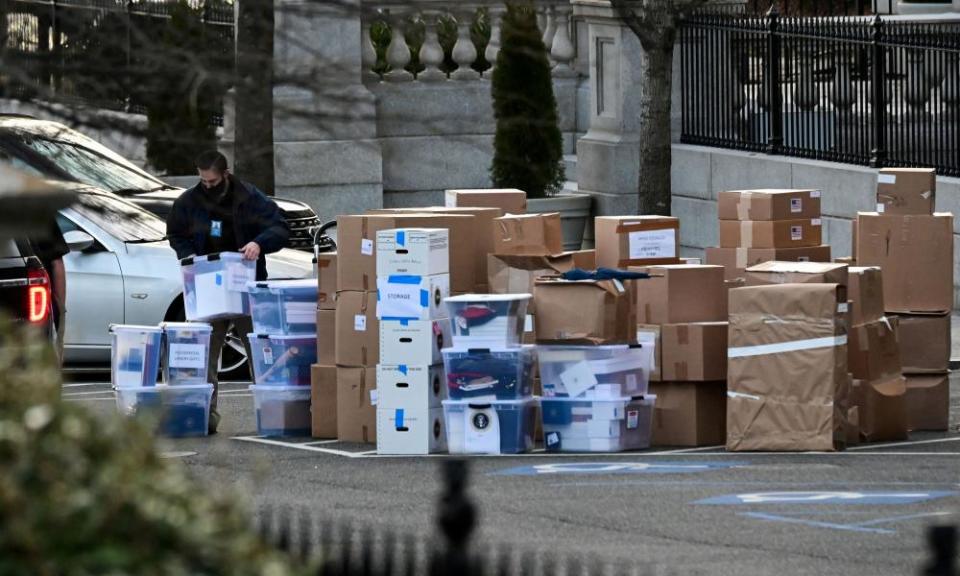 A worker in a dark jacket standing in a parking area lifts a clear plastic bin filled with documents from several large piles of bins, boxes and large rectangular objects wrapped in brown paper.