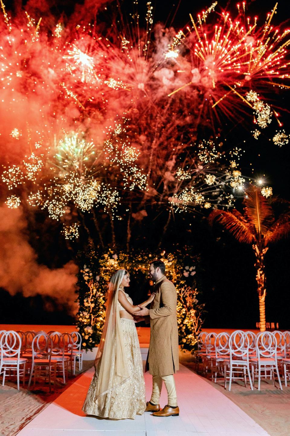 A bride and groom hold hands as fireworks explode above them.