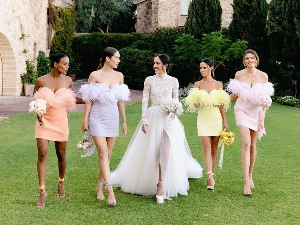 A bride stands surrounded by bridesmaids in brightly colored mini dresses trimmed with feathers.