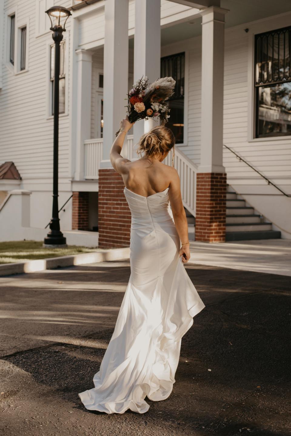 A bride walks towards a house and raises her bouquet in the air.