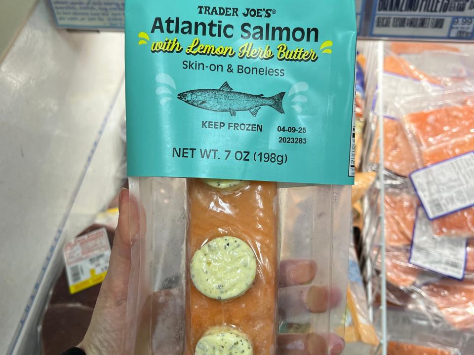 A hand holding a package of skin-on, boneless Trader Joe's Atlantic salmon with lemon-herb butter.