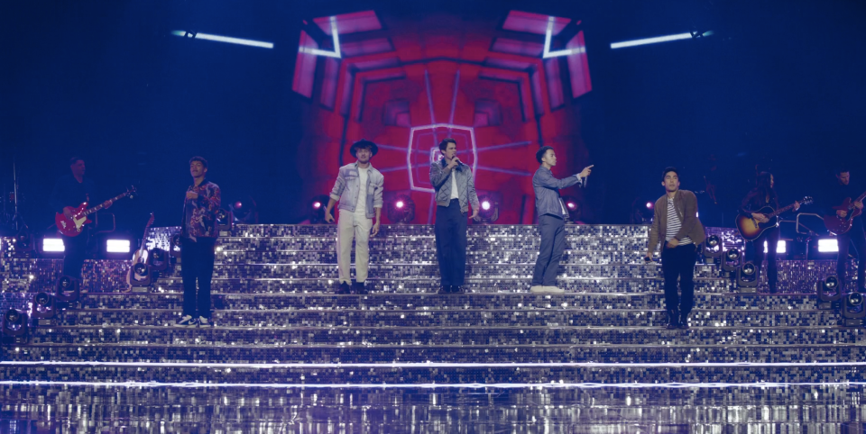 Band performs on stage with lit-up steps, members in stylish suits, dynamic poses, and energetic expression