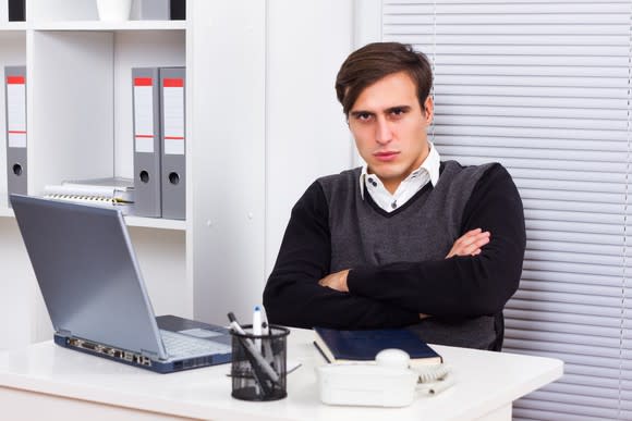 An angry young worker sitting in front of laptop with his arms crossed.