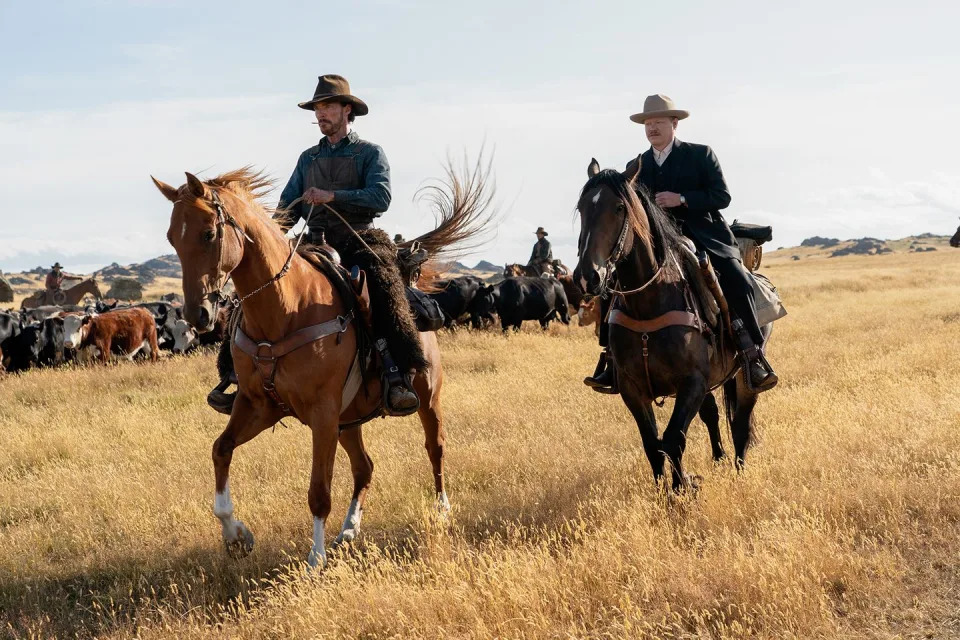 benedict cumberbatch and jesse plemons in character for the power of the dog, both men ride brown horses in a field with cattle behind them and have on hats