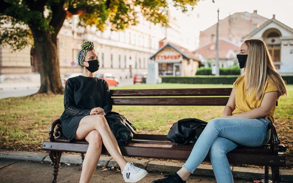 Two women with black face masks sitting on bench in park