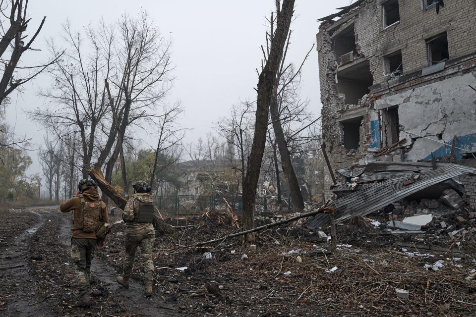 two people, on in camo, both wearing helmets, walk on dirt road by destroyed 4-story building