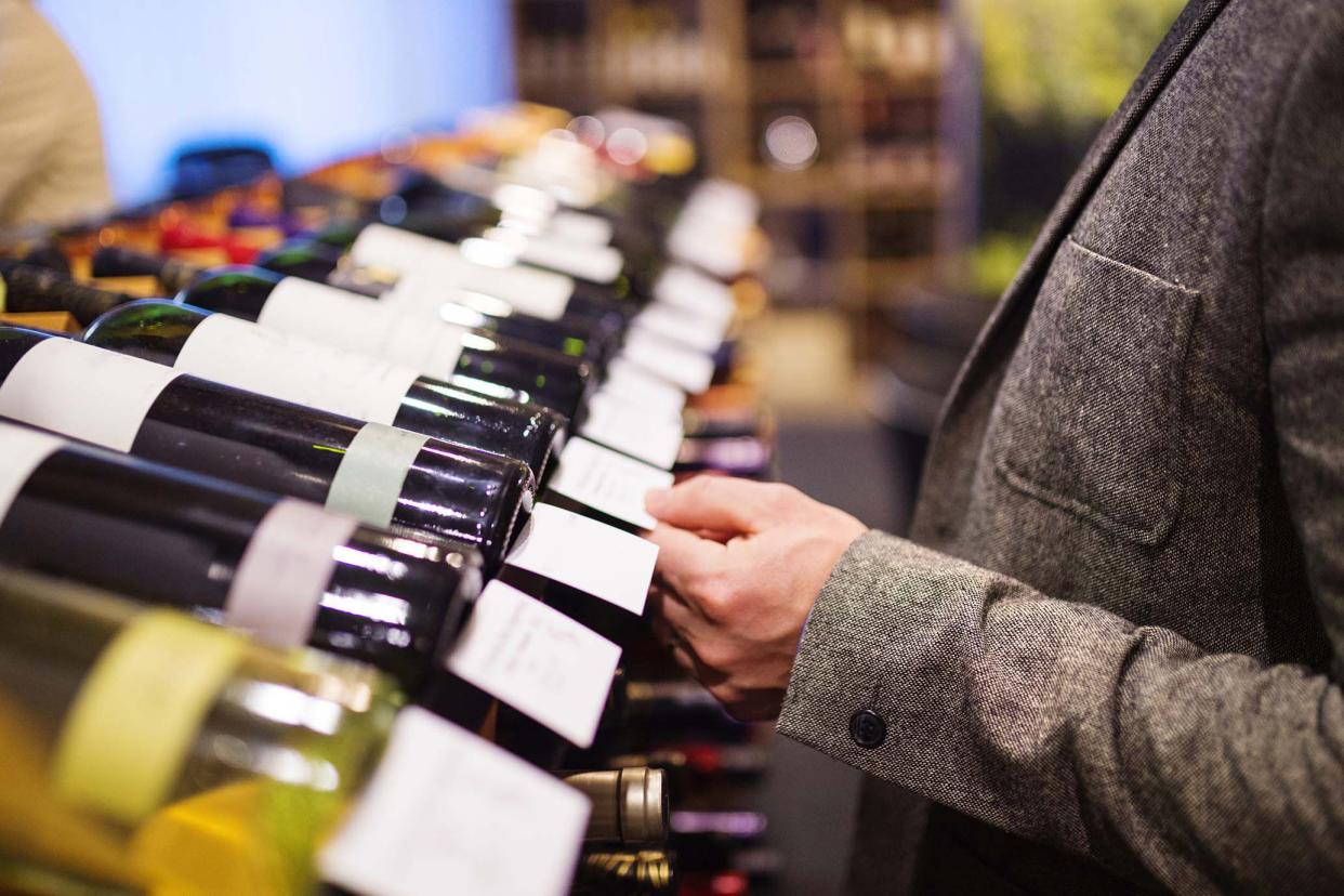 Mid-section of a man wearing an overcoat inspecting wine descriptions next to wine bottles along a row of wines in a store, with a blurred background of shop and other wines for sale