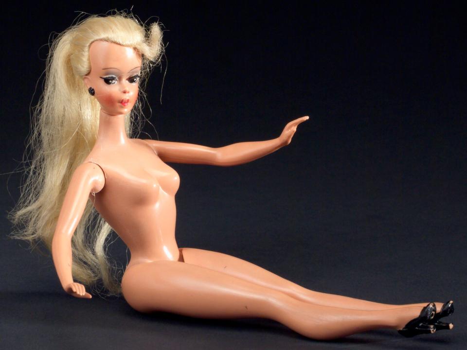 Blonde Bild Lilli doll, unclothed, made of all-rigid plastic and jointed limbs. Pictured sitting down with a plain black background