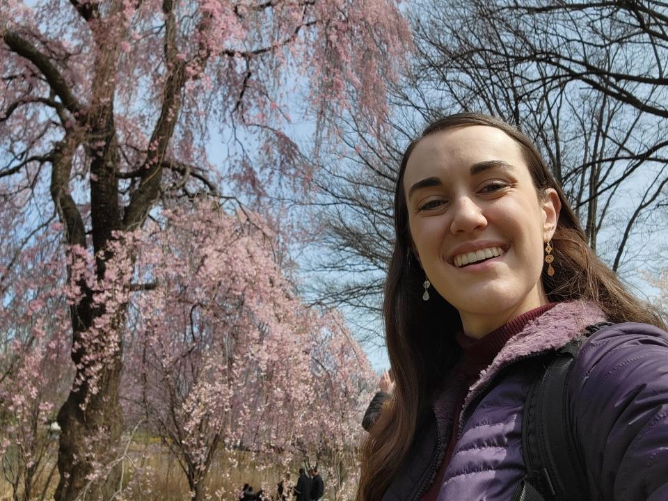 Amanda smiles in front of a tall tree with draped, pink-blossomed branches.