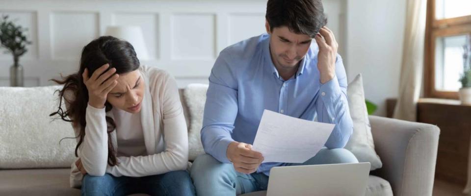 Anxious young couple look at bills while sitting on couch, holding heads in hands