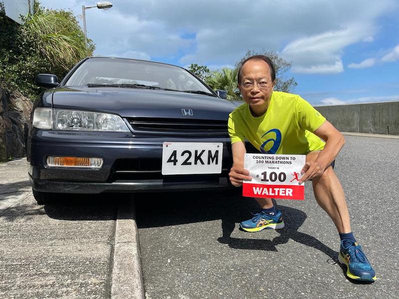 完成第一百個馬拉松的張樹槐，在其車前手持跑友特製的號碼布，車牌正是全馬路程42KM。