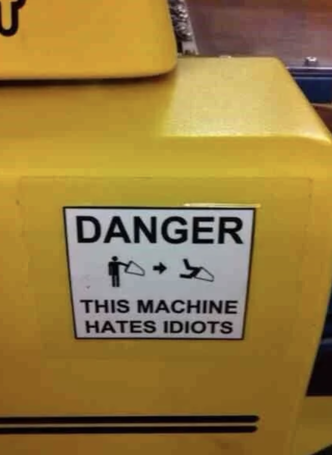 Warning sign on machinery humorously reading "DANGER - THIS MACHINE HATES IDIOTS," with pictograms illustrating harm