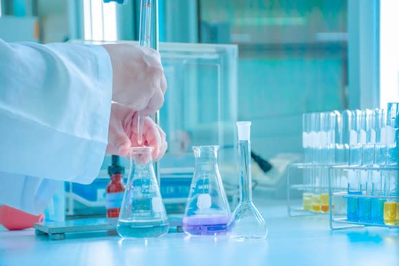 A lab worker's hands holding a beaker with some liquid in it that's sitting on a table along with other beakers and test tubes.