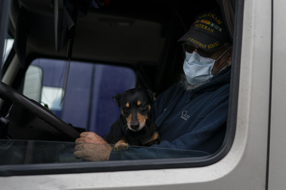 In this April 5, 2020, photo, Ronnie Jackson, of Earlsboro, Okla., wears as face mask in the cab of his semitruck with his dog Shorty on his lap before rolling west from the TA Travel Center truck stop in Foristell, Mo. Jackson wears a face mask to protect himself and others during Coronavirus outbreak. (AP Photo/Carolyn Kaster)