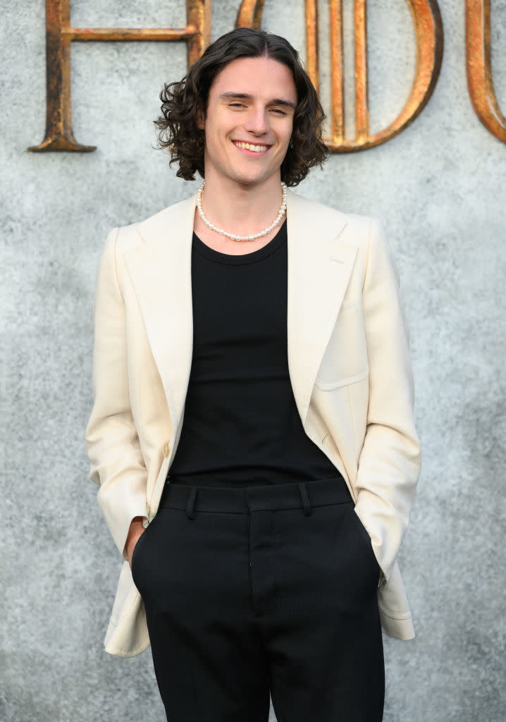 Smiling man with shoulder-length curly hair wearing a black shirt, cream-colored blazer, black pants, and a pearl necklace. Background is blurred