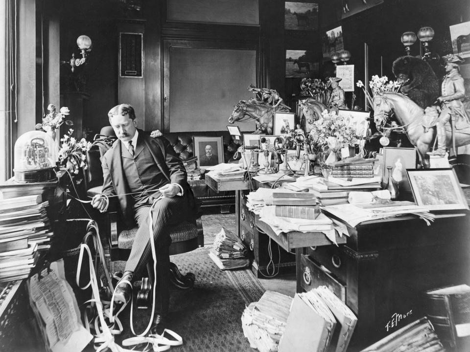A stockbroker surrounded by books, papers, and figurines in his office in 1904.