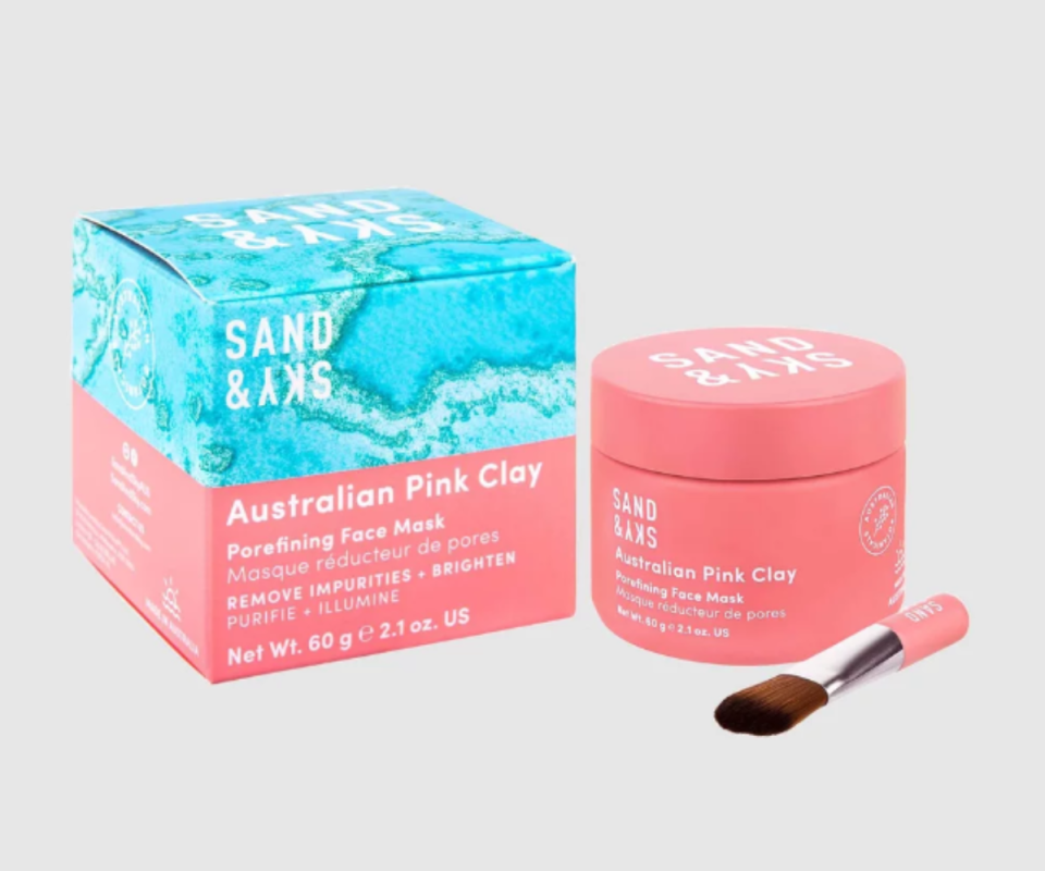 A pink tub of Sand & Sky's pink clay mask sits on the right against a pale grey background with an application brush in front of it with the packaging box on the left in pink and blue.