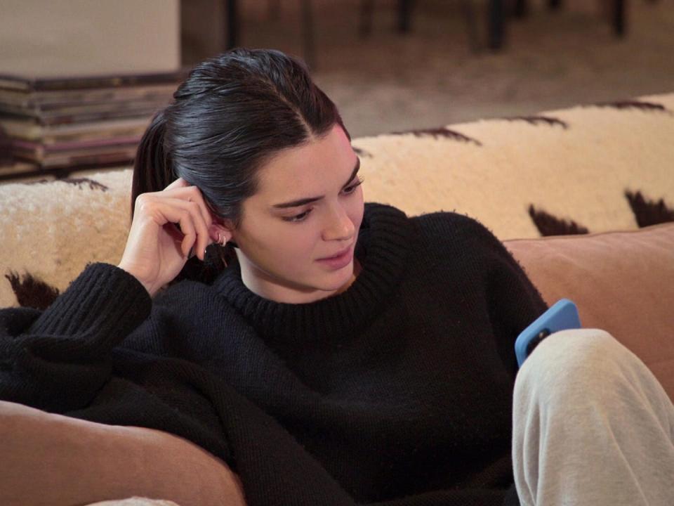 kendall jenner sitting on a couch looking at her phone, resting her had on her hand and leaning against the arm rest