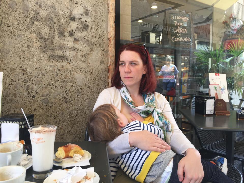 Chrissy Fleishman, a white woman with red hair, breastfeeds her young child while sitting at a restaurant table.