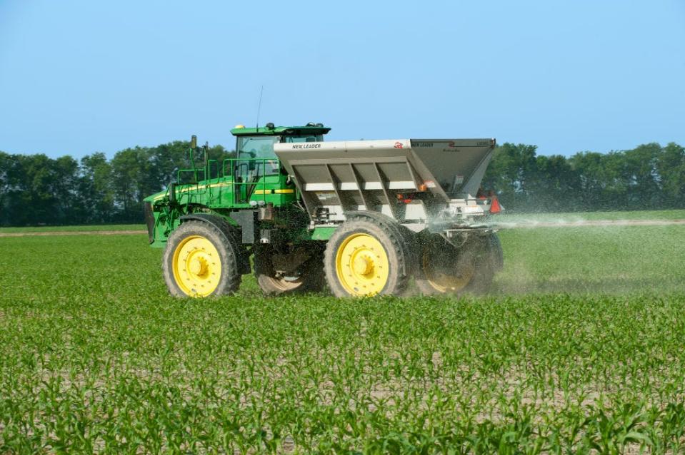 A John Deere Hi-Boy with a New Leader Spin Spreader Box applies dry pelleted nitrogen fertilizer to an early growth grain corn crop in the spring.