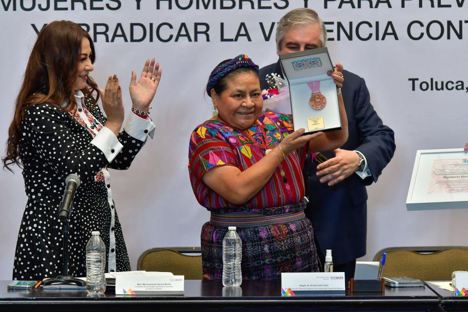 rigoberta menchu smiles as she holds up a medal in a box, she wears a multicolored headband, blouse, and skirt