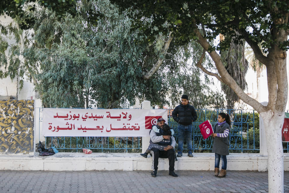 A Tunisian man and his family gather next to a banner celebrating the anniversary of Tunisian revolution, in the city of Sidi Bouzid, Friday, Dec. 17, 2021. Banner in Arabic reads: "Sidi Bouzi celebrates the anniversary of the revolution." President Kais Saied changed the anniversary date of Tunisia's 2011 revolution – when former autocratic ruler Zine el Abidine Ben Ali fled the country – to Dec. 17 to mark the day in 2010 when fruit seller Mohammed Bouazzi set himself alight, setting off the series of uprisings in Tunisia that led to what is now known as the Arab Spring. (AP Photo/Riadh Dridi)