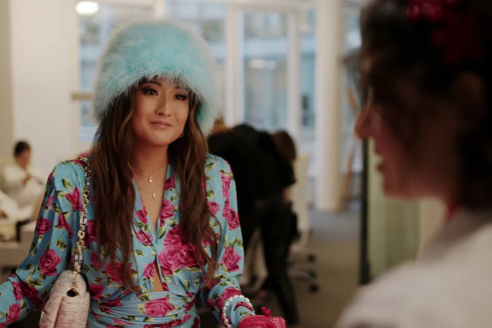 Ashley Park smiling in a floral dress and furry hat while holding a handbag, talking to another person in "Emily in Paris"