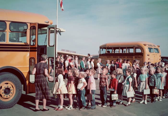 Children in 1960s attire, including dresses and pants, line up to board a yellow school bus outside of Justo Comolak School in the Marysville Unified School District