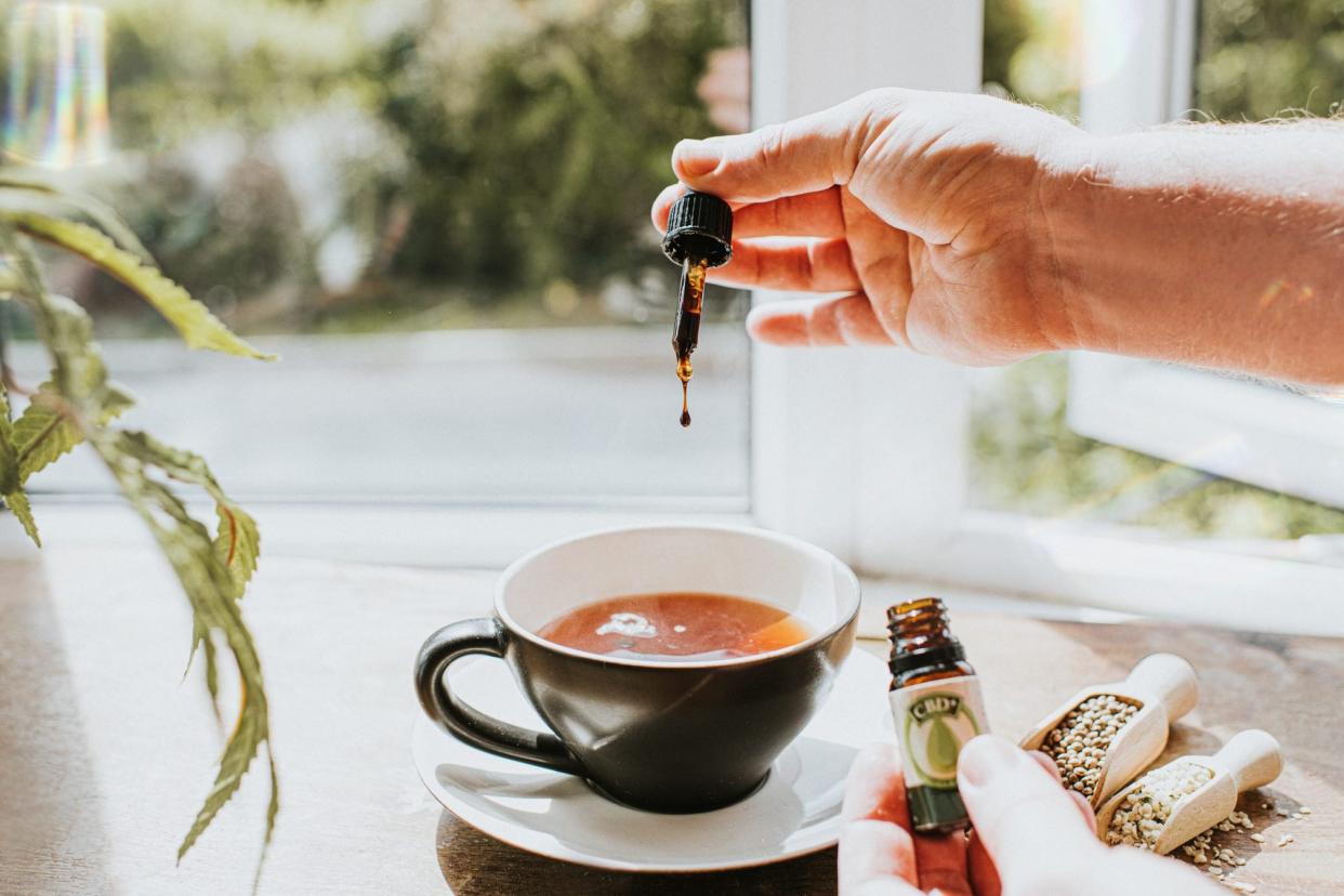 Hand dropping CBD oil into a Cup of Tea, surrounded by Cannabis Plants