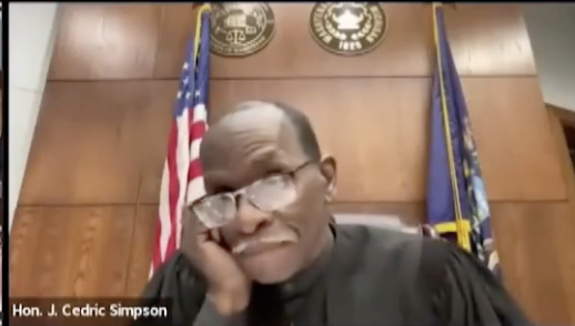 Hon. J. Cedric Simpson in judge's robes, sitting in front of a wood-paneled wall with an American flag and state flag, looking contemplative